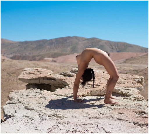 flexible naked woman standing in a backward arch on all four hands and feet. She's on a promontory surrounded by dropoffs into a canyon with scenic views in all directions.