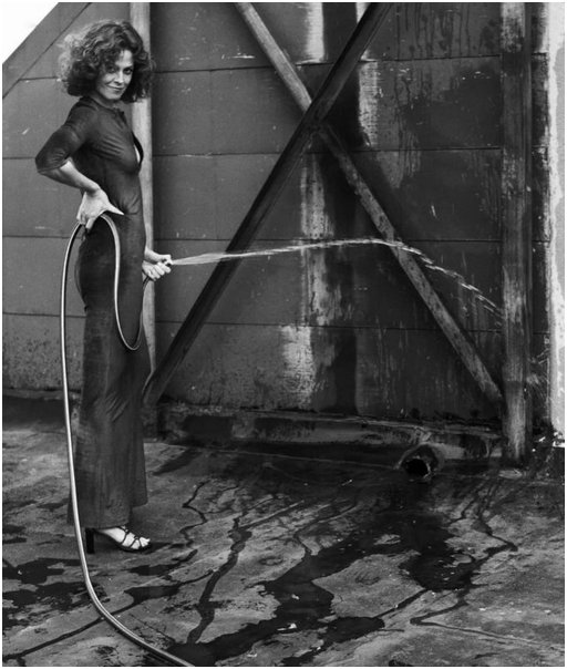 Sigourney Weaver in a slinky dress holds a garden hose as if it were her penis and she was peeing